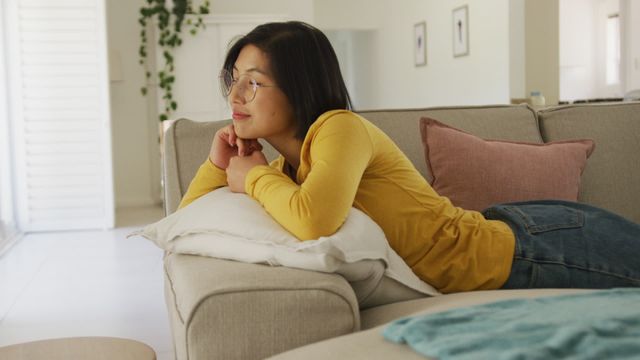 Asian woman enjoying her time at home, lying on couch with head resting on a pillow. Perfect for use in home lifestyle blogs, leisure time concepts, comfortable living advertisements, and content related to modern home interiors.