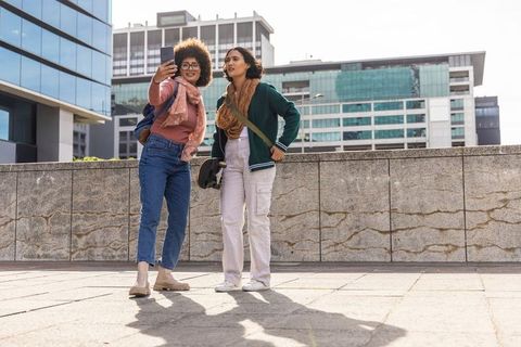 Two friends taking selfie on sunlit urban plaza with glass office towers and casual street style