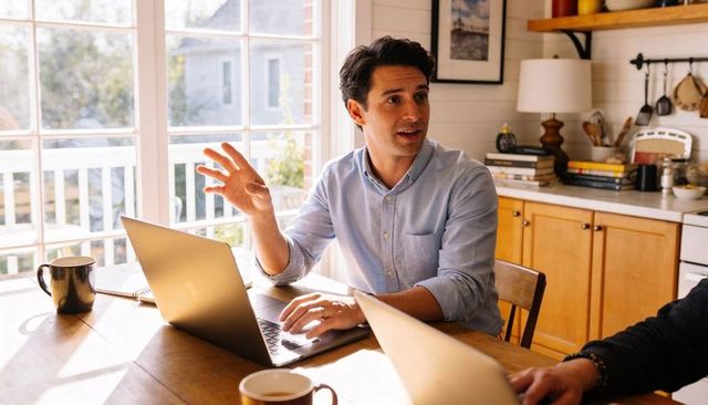 Man discussing remote work while working from home kitchen table with laptop and coffee