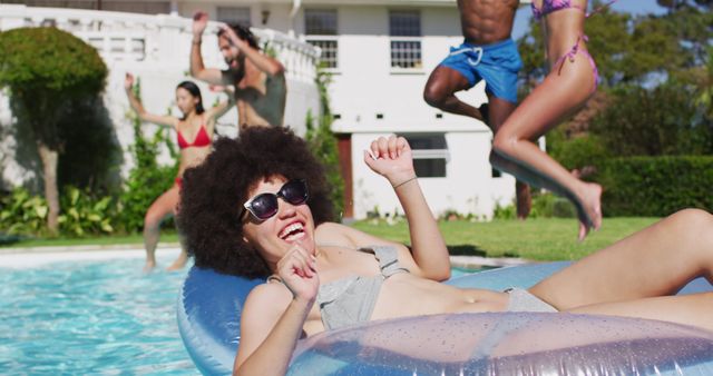 Group of friends having fun at a pool party while a woman in a bikini is relaxing on an inflatable float. One friend is smiling, relaxed, and wearing sunglasses. Ideal for promoting summer activities, pool parties, leisure time, vacation packages, and fun outdoor gatherings.