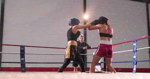 Female boxers sparring in ring under spotlight, coach observing intense training