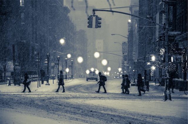 People are braving heavy snowfall on a busy urban street at night. The scene includes prominent traffic lights and city illuminations, giving it a cozy yet chilly atmosphere. Ideal for projects depicting city life in winter, winter travel, urban lights, and night-time city scenes.