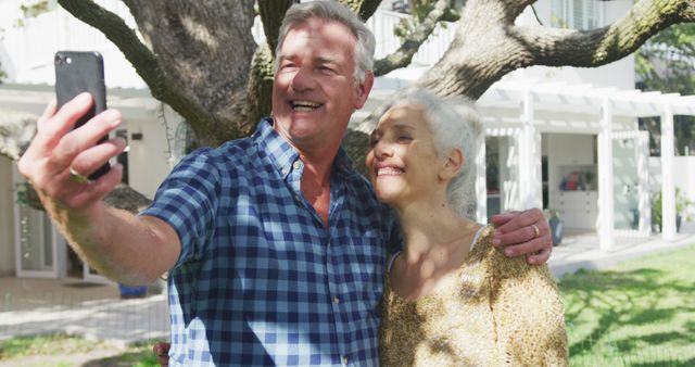 A joyful older couple stands in their backyard, holding a smartphone, and taking a selfie together. They are smiling and embracing, with greenery and a large tree as their backdrop. This image is great for illustrating themes of happy retirement, senior lifestyle, love in older age, and enjoying life outdoors. Perfect for use in promotional materials, senior lifestyle blogs, and advertisements for outdoor or leisure products.