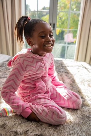 African american girl sitting on bed wearing pink onesie smiling in sunlit bedroom