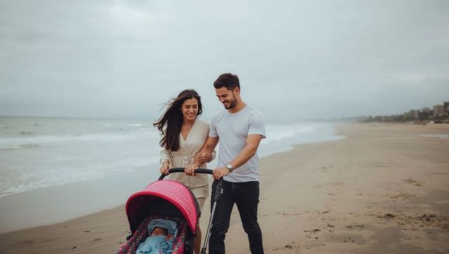 Smiling couple walking and pushing pink stroller along ocean shore with sleeping infant