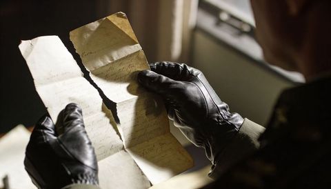 Uniformed man wearing black gloves examining torn handwritten letter under window light