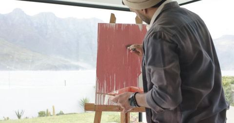 Indian male painter working on dripping red canvas at easel by window with mountain view