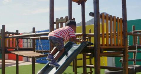 Child climbing up slide on colorful wooden playground with rope bridge and turf