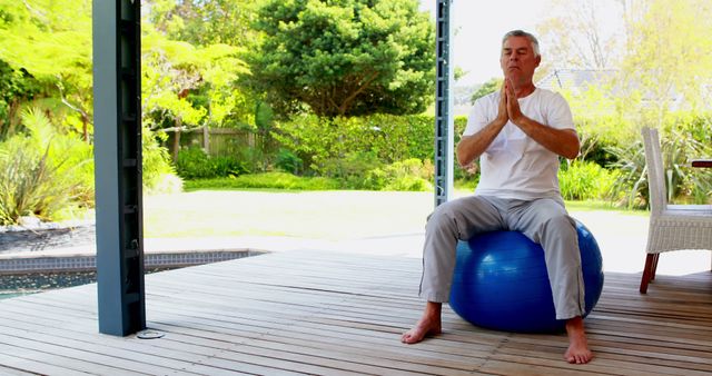A middle-aged Caucasian man practices yoga on a blue exercise ball on a wooden deck, with copy space. His eyes are closed in concentration, and his hands are pressed together in a meditative pose, promoting health and relaxation.