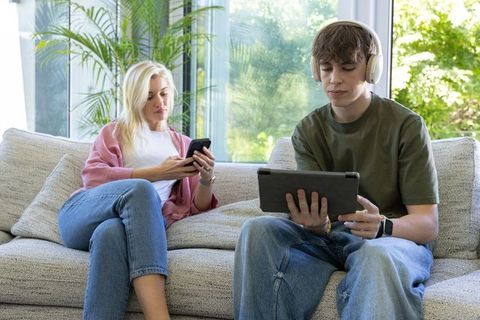 Adult woman and teenage boy relaxing on sofa using smartphone and tablet at home