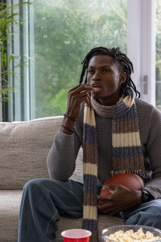 African american man watching game eating popcorn holding football on couch