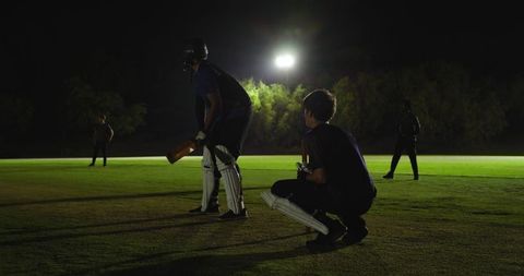 Night cricket practice under floodlights: batsman and wicket-keeper training on grass pitch