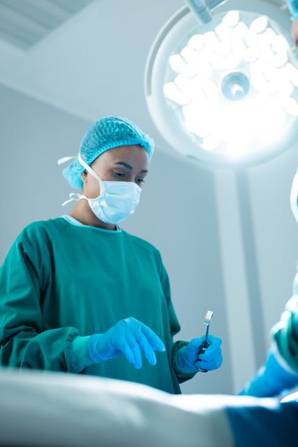 Biracial female surgeon wearing face mask and gloves, holding surgical instrument during a procedure in a well-lit operating room. Ideal for use in medical and healthcare-related content, hospital brochures, educational materials, and articles about surgical procedures and healthcare professionals.