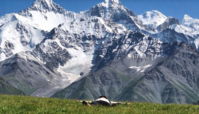 Man lying with arms out on alpine meadow under towering snow-capped mountain peaks