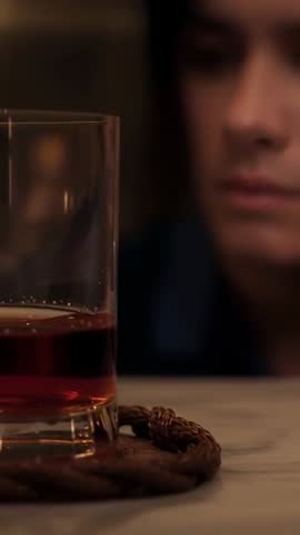 Vertical lounge video of man musing at bar while camera racks to amber tumbler closeup