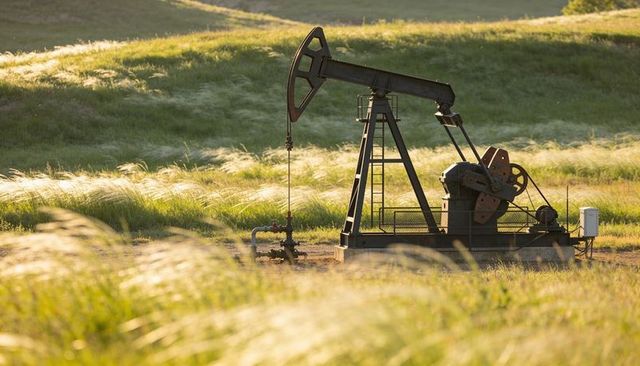 Pumpjack operating in backlit rolling grassland at golden hour on fenced concrete well pad