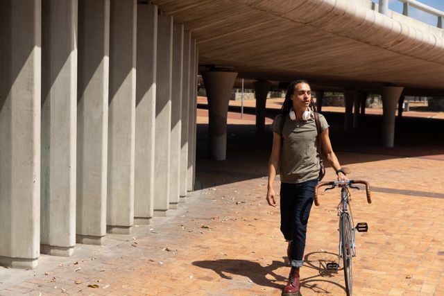 Biracial alternative man with dreadlocks out and about in the city on a sunny day, walking with his bike next to bridge. Urban trendy man on the go.