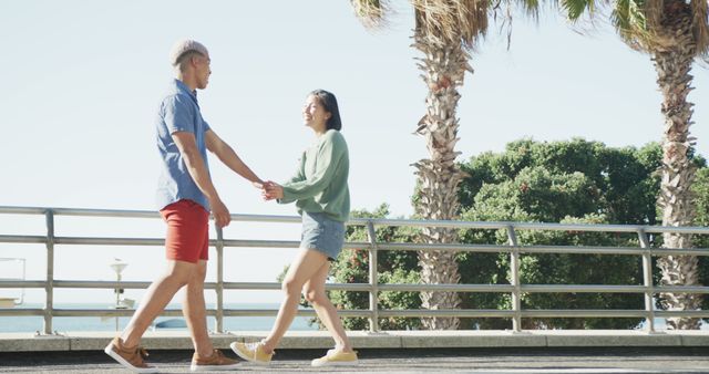 Happy diverse couple walking and holding hands on sunny promenade by the sea. Summer, vacations, romance, relationship and free time, unaltered.