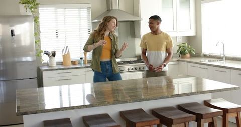 Couple chatting and laughing in modern kitchen with large stone island and wooden stools