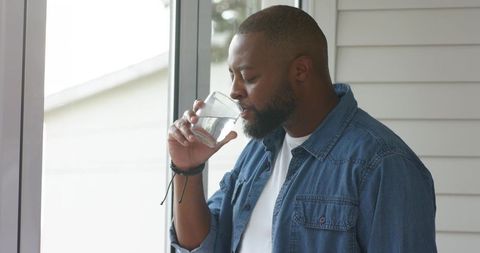 Man sipping water by sunlit window in denim shirt for calm casual wellness lifestyle