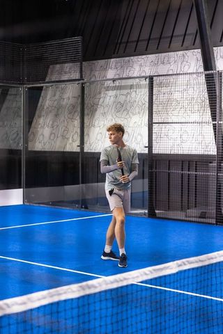 Male padel player gripping racket on vibrant blue indoor court with glass mural backdrop