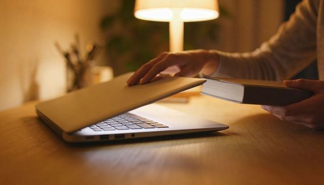Closing laptop while holding book at cozy wooden desk under warm lamp light