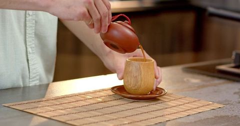 Pouring tea from ceramic teapot into wooden cup on bamboo mat