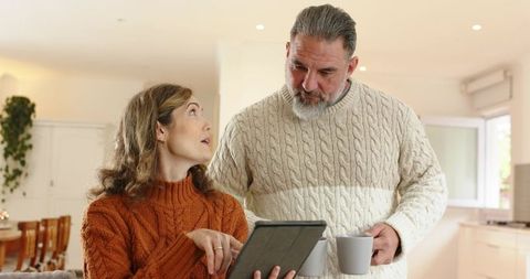Couple Discussing Tablet in Minimalist Kitchen Wearing Cable Knit Sweaters Holding Mugs