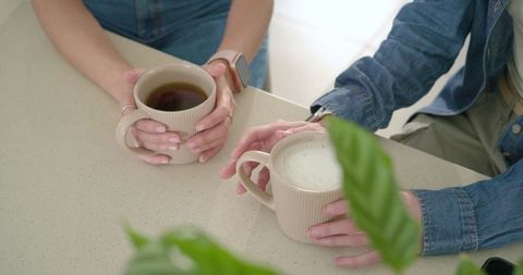 Two friends holding ribbed beige mugs on kitchen counter, smartwatch and ring visible