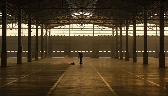 Silhouetted worker sweeping dust across empty industrial warehouse floor at sunrise
