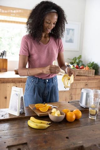 African american woman peeling banana preparing fresh smoothie with oranges in sunlit kitchen