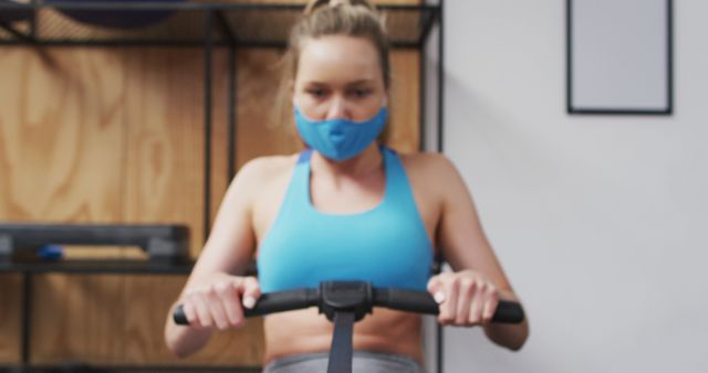 Photo depicts a woman in sports attire exercising on a rowing machine in a gym while wearing a face mask for protection. This visual is useful for health and fitness blogs, workout tips, gym promotions, and articles about maintaining fitness routines during a pandemic.