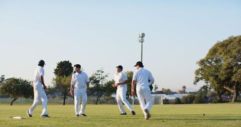 Diverse male cricket players walking on sunny outfield wearing white uniforms and caps