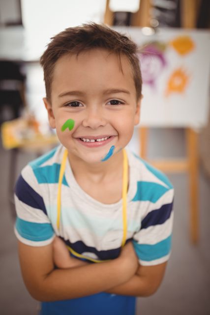Young boy smiling with paint on his face, standing in an art class. Ideal for educational materials, art class promotions, and creative learning content.