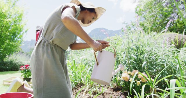 This image depicts a woman engrossed in gardening activities under bright sunlight. She wears a light dress and a wide-brimmed hat while carefully watering plants and flowers. The lush green garden and vibrant flowers suggest a beautiful summer day. This picture can be ideal for use in articles or advertisements related to gardening, outdoor activities, summer leisure, horticulture tips, or lifestyle blogs. It could also illustrate content on sustainable living, nature conservation, and plant care tutorials.