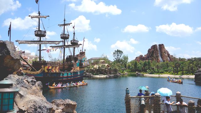 Tourists and families are seen enjoying various outdoor activities at a theme park with a pirate ship and lake. Ideal for promotions related to vacations, family outings, amusement parks, and outdoor summer fun.