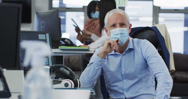 Senior businessman wearing a face mask while working at his desk in a modern office. Ideal for topics on corporate life during a pandemic, health and safety protocols at work, modern office environments, and professional management scenarios.