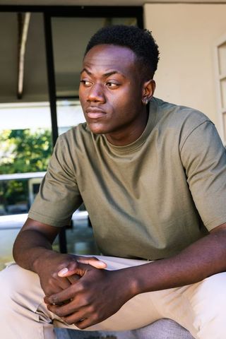 Young man sitting on porch gazing through glass door wearing olive tee and beige pants