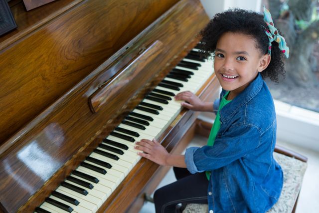 Young girl with curly hair smiling while playing the piano at home. She is wearing a denim jacket and a colorful bow, showing great joy and enthusiasm. This image can be used for educational content, music lessons, advertisements for children's activities, or home life scenes.