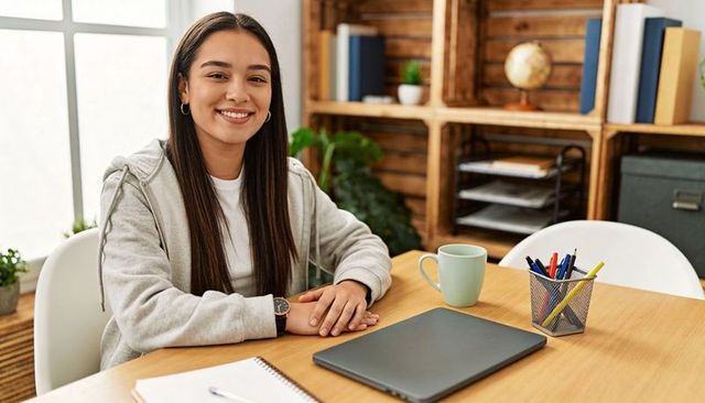 Smiling young woman in gray hoodie at minimalist home office desk with laptop and plants