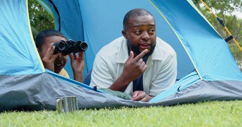 African american father and teen son camping, pointing and peering with binoculars outdoors