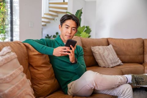 Young asian man relaxing on leather couch using smartphone in bright modern living room