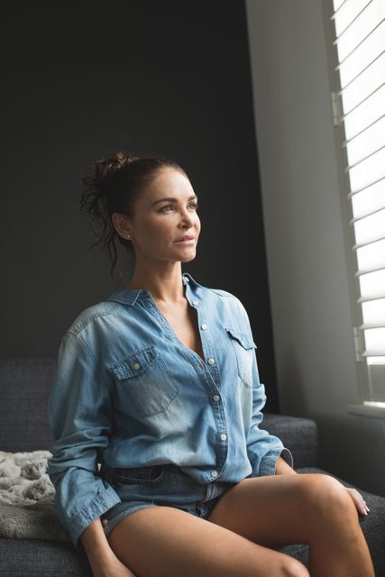 Woman sitting on sofa in a comfortable home, wearing a denim shirt and looking out the window. Natural light illuminates her face, creating a serene and thoughtful atmosphere. Ideal for use in lifestyle blogs, home decor magazines, mental wellness articles, and advertisements promoting comfortable living spaces.