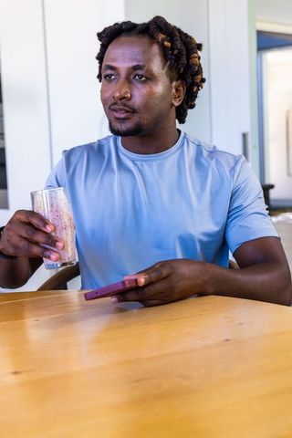 African american man drinking smoothie and checking phone at sunlit modern kitchen table