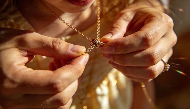 Woman fastening gold knot necklace closeup in warm sunlight with gemstone ring