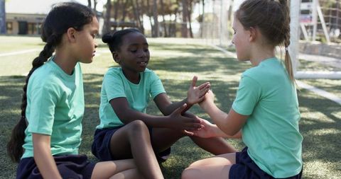 Diverse school girls playing hand-clap game on soccer field wearing green shirts and navy shorts