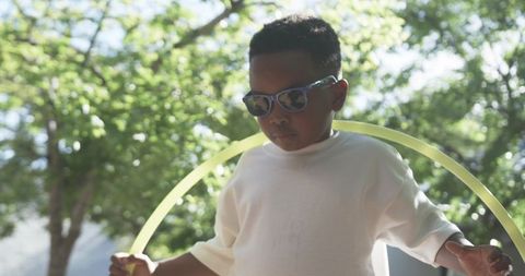 Boy holding neon hula hoop in sunlit park wearing sunglasses and light long-sleeve shirt