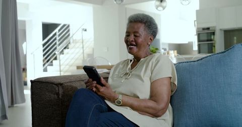Senior woman smiling while using smartphone on modern sofa in contemporary living room