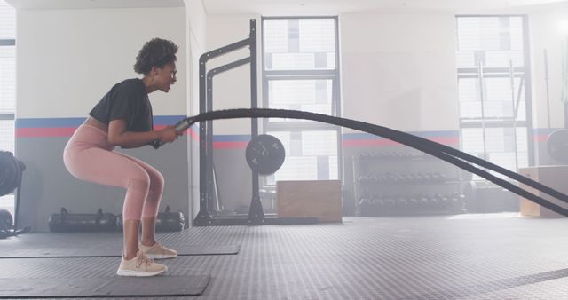 Fit young woman exercising with battle ropes in modern gym. She looks determined while engaging in an intense workout, emphasizing strength and fitness. Ideal for use in health, fitness, and lifestyle websites to convey active and healthy lifestyles, promoting gym equipment, fitness programs, or athletic wear.