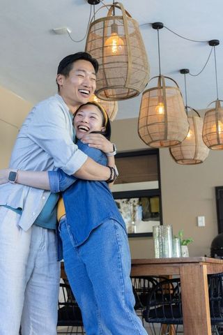 Asian couple embracing and laughing in cozy dining room with rattan pendant lights